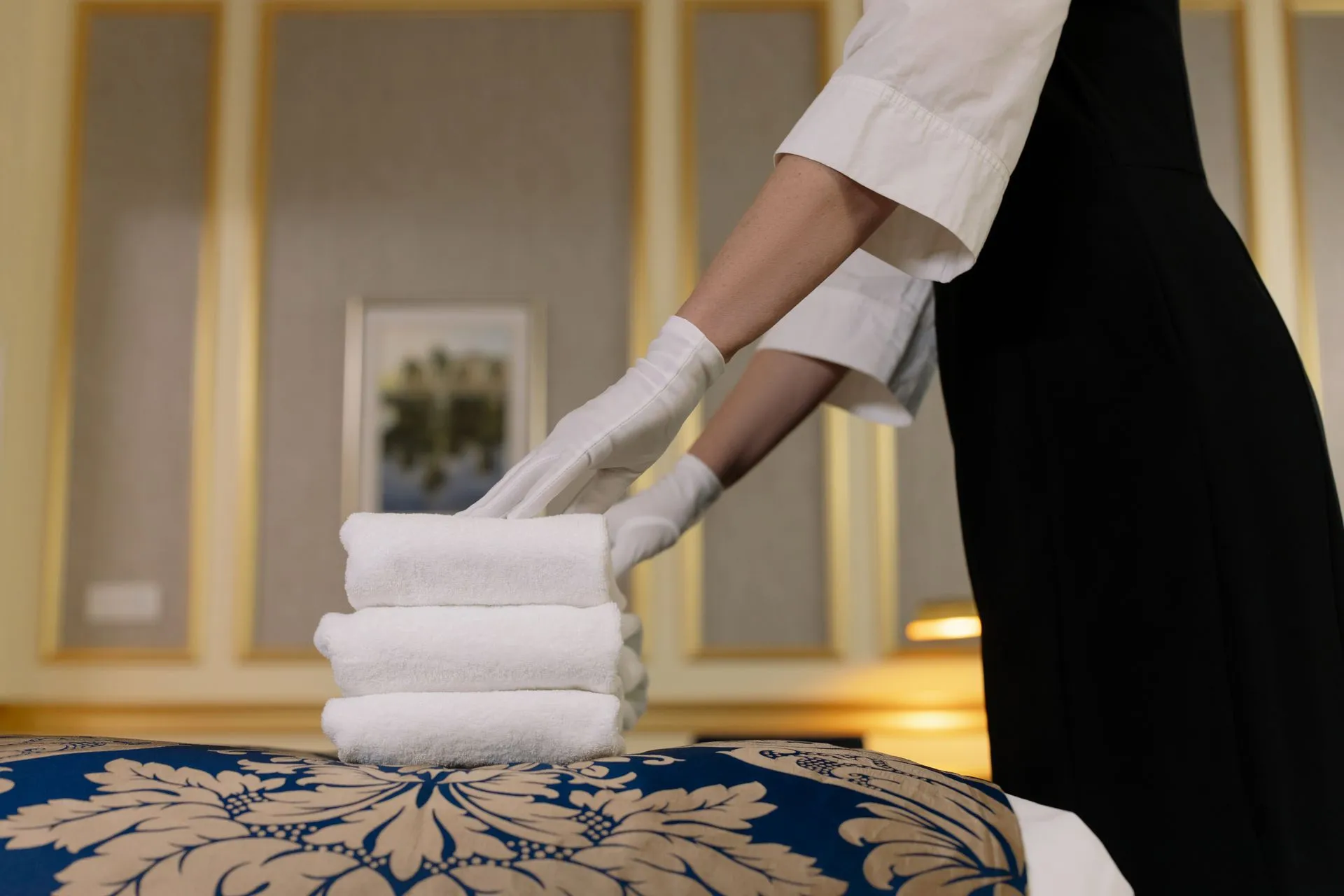 A housekeeper in white gloves placing a stack of folded towels on a hotel bed